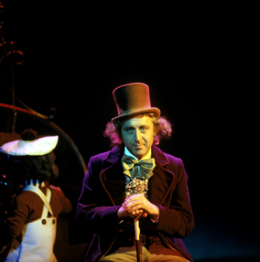 A man in a purple suit and a brown top hat with a cane is sitting down next to a worker.